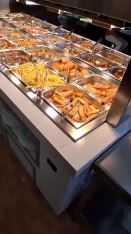 The video pans across a buffet with various food options. In the foreground, there are trays of french fries, spring rolls, and fried chicken. Further back, more trays hold a variety of dishes, including what appear to be meats, vegetables, and breaded items. In the background, several people are visible, some appearing to be customers selecting food, and others working. The setting is likely a restaurant or buffet establishment. The lighting suggests it could be daytime or a well-lit indoor space. There are no overt emotional cues from the people in the video. No visible text is present.