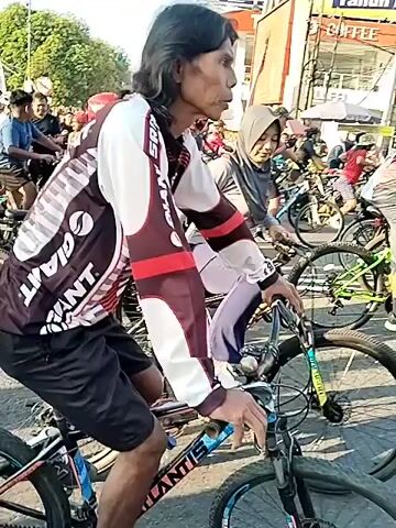 A large group of people, primarily adults, are depicted outdoors engaged in a cycling event. Many individuals are on bicycles, which appear to be mountain bikes or hybrid models. Some cyclists wear helmets, and several women are seen wearing hijabs. One prominent cyclist in the foreground wears a black and white jersey with "GIANT" text. The video shows participants both actively cycling on a paved road and gathered in queues, seemingly waiting at a starting line or checkpoint. A large blue inflatable archway, bearing the text "ELITE CELL," is visible in the background of a segment. The environment is sunny and bright, with buildings and other urban elements present in the distant background.