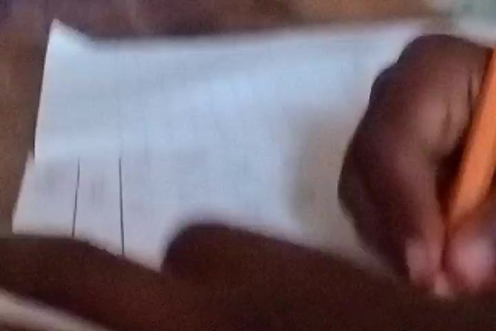 A hand holding an orange pencil is writing on a piece of paper. The hand is brown-skinned. A blue cloth obscures much of the scene behind the hand. Stacks of white buckets can be seen in the background. There is some indiscernible writing already on the paper.
