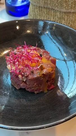 A plate of steak tartare topped with sprouts and a raw egg yolk is centered on a dark bowl. The bowl is on a table with a basket of bread, french fries, a pot of mussels, and drinks.