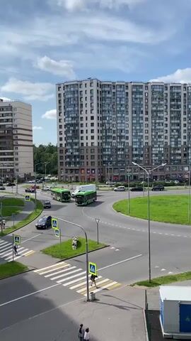 The video captures a street intersection with heavy pedestrian and vehicle traffic. Two green buses are prominently featured, one appearing to be a trolleybus due to overhead wires. They navigate a roundabout alongside other vehicles, including cars and a white truck. Pedestrians are seen crossing at marked crosswalks, some walking alone, others in pairs. The environment includes modern high-rise apartment buildings, streetlights, traffic signs, and green spaces with grass. The sky is partly cloudy. The overall scene depicts a busy urban setting.