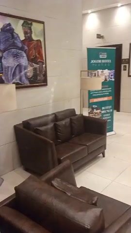 The video shows the interior of a hotel lobby in Ibadan, Nigeria. The scene is brightly lit, suggesting it is daytime. The lobby features comfortable seating areas with brown leather sofas, and artwork on the walls. One painting depicts three figures in traditional attire, possibly representing a cultural or historical scene. Another banner, partially visible, advertises "JOGODOR HOTEL IBADAN," indicating the hotel's name and location. The overall atmosphere appears calm and inviting, suitable for a hotel reception area. The flooring is tiled, and there are lamps and potted plants adding to the decor. The camera pans to reveal a corridor leading deeper into the hotel, with more artwork and an air conditioning unit visible on the wall. The video ends with a glimpse of a doorway or entrance, with some lights visible in the distance.