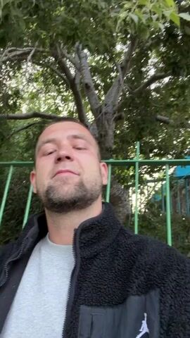A man takes a selfie outdoors. He has a short haircut, a beard, and is wearing a gray shirt under a black jacket with a white logo on the pocket. Behind him are green trees and a green fence. The man smiles slightly at the camera. The camera angle is low, looking up at the man, and he briefly looks up towards the sky.