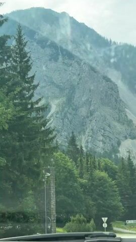 This video shows a view from inside a vehicle moving along a road, looking out at a steep, rocky mountainside. The mountain is characterized by a large scree slope, with debris from rockfalls clearly visible. Patches of green vegetation, including coniferous trees and deciduous trees, are present on the lower slopes and in the foreground. In the lower left of the frame, a stone structure resembling a monument or pylon stands beside the road. Traffic signs are also visible on the right side of the road. The sky is overcast. The camera appears to be looking slightly upwards towards the mountain.