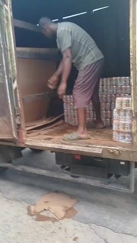 The video shows a person in a truck, unloading cans of beverages. The truck appears to be parked on a street, possibly in Yenagoa, Nigeria, given the location context. The person, wearing a green shirt and patterned shorts, is seen handling stacks of canned drinks. The truck's cargo area is visible, with a large, wooden bin-like structure present. The scene appears to be outdoors, with daylight suggesting it's daytime. The ground is paved with concrete.

A notable detail is that one of the stacks of cans is being thrown out of the truck towards the viewer. This action is captured from a different angle in the latter part of the video, where the cans are mid-air, being caught by someone off-camera. The overall impression is one of a delivery or distribution process, possibly a quick or informal one. There is no visible text in the media. The weather appears to be clear.
