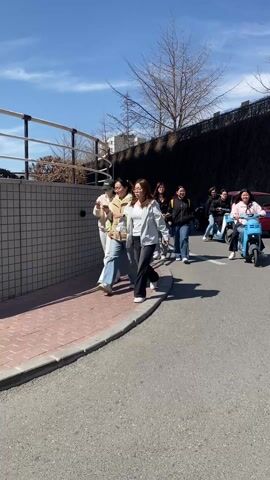 The video shows a group of people walking down a sidewalk next to a road. Some people are riding light blue electric scooters in the same direction as the walking group. The road and sidewalk are paved, and there's a short brick wall separating them. On the left is a wall with a metal railing. On the right, there is a tall retaining wall and some trees without leaves. The sky is bright blue with a few clouds. The people appear to be young adults or college students. The overall impression is a sunny day on a college campus.