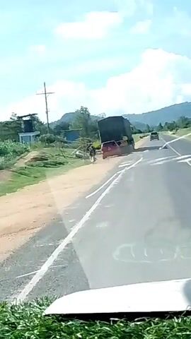 A video shows a red vehicle with a dark load in its bed pulled off to the side of a paved road. There is a person walking next to the vehicle on an unpaved portion of the road, near a utility pole and some small buildings. In the distance there are small mountains. The video appears to be taken from a moving car as it passes the parked vehicle.