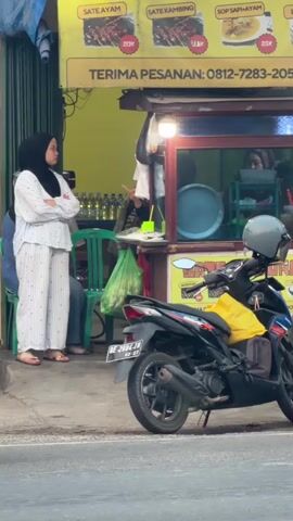 A woman wearing a white hijab and a matching white outfit with pinstripes stands with her arms crossed outside a food stall. The stall has a yellow awning displaying pictures of food and text in Indonesian, including "TERIMA PESANAN: 0812-7283-205". Behind the woman, several green chairs are visible. A motorcycle with a yellow bag on the back is parked in front of the stall. The street is paved, and there are signs of traffic blur, indicating movement. The woman appears to be waiting.