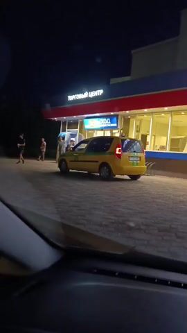 A yellow car with green stripes and a taxi sign on its roof is parked in front of a building at night. The building has signs that read "ТОРГОВЫЙ ЦЕНТР" and "SHERIFF". There are at least three people standing near the car and the building. The ground is paved with bricks. The scene is captured from inside another vehicle.