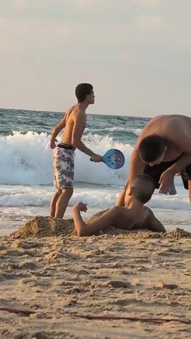 At 00:00, three young males are on a sandy beach near the ocean. The waves are breaking on the shore. One male in the foreground is lying partially buried in the sand, resting on his forearms. Another male stands behind him, reaching towards the first male with his right hand. A third male stands to the left, also facing the others, with his right arm extended. He is holding a small, blue paddle. At 00:01, the male in the middle shifts his weight. At 00:02, the standing male to the left is still holding the paddle. At 00:03, the male buried in the sand begins to sit up slightly, raising his left arm. At 00:04, the standing male to the left moves closer to the boy in the sand, holding the paddle out towards him. Another person can be seen in the water behind the breaking waves.