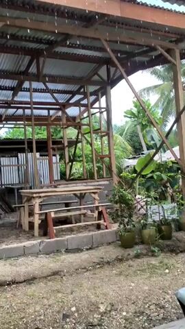 A light rain falls on an unfinished structure with a corrugated metal roof and a wooden frame. A wooden table and benches sit inside the structure. Trees and potted plants are visible in the background. The ground is covered with dirt and gravel.