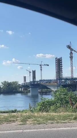 A video recorded from a moving vehicle captures a large bridge under construction over a wide river on a sunny day. Multiple tall tower cranes are visible around the unfinished concrete and steel pylons of the bridge, which appears to be a cable-stayed or suspension design. A central support pier stands in the river. A small boat with a person is seen on the water in the distance. The foreground consists of green grass and a paved area, while lush green bushes and trees line the riverbanks. The sky is bright blue with scattered white clouds.