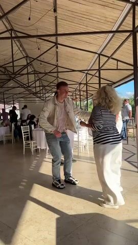 A group of people are dancing under a covered outdoor pavilion. The floor appears to be tiled. The ceiling is made of wooden planks and metal beams. Sunlight streams in from the right, casting shadows on the floor. In the foreground, a man in a light-colored jacket, striped t-shirt, and jeans is dancing. To his right, a woman with blonde, curly hair, wearing a striped shirt and white pants, is also dancing. Behind them, other people are visible, some dancing and others seated at tables in the background. The overall atmosphere suggests a casual gathering or party.