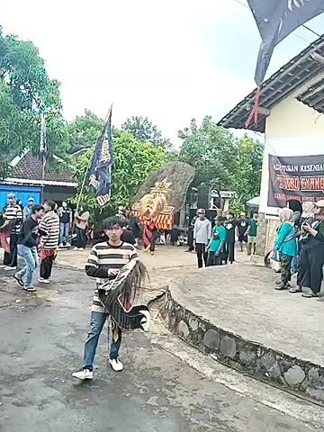The video shows a cultural event in a village setting. Several people are present, with some wearing striped shirts and carrying fringed objects. A large, elaborate figure with orange and yellow details is being moved through the crowd. A flag with writing is also visible. The scene takes place outdoors, with trees and buildings in the background. The sky is overcast.