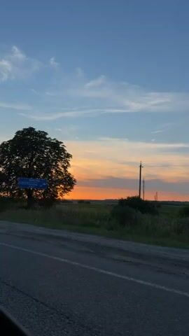 The video shows a car's perspective driving along a road at sunset. The sky is a mix of blue, orange, and pink hues. There are trees and fields on the side of the road. In the first few seconds, a signpost with Cyrillic text is visible next to a large tree. Later, a white semi-trailer with the logo "CGVL" is seen. The video concludes with a wide view of the sunset over the horizon, with power lines and trees silhouetted against the colorful sky.