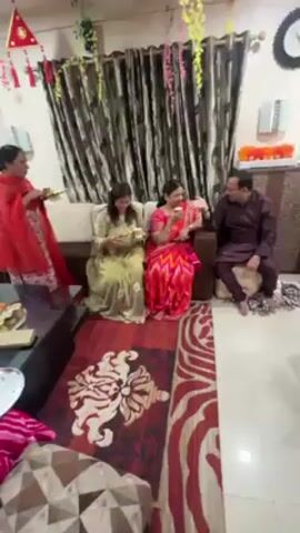 The interior of a decorated room shows a group of women in traditional clothing, many holding plates of food. Some women are in bright saris, others in more subdued shades.  A man in a maroon outfit sits on a cushion on the floor.  The room is decorated with festive garlands and a decorative kite. A patterned rug covers the floor. The room seems to be set up for a celebration or gathering. The women appear to be taking a break and having a snack. They are talking amongst themselves.