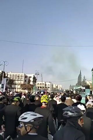 A large group of people, many wearing helmets, are gathered on a street in what appears to be a city. A paraglider is visible in the sky. Green smoke or haze drifts in the air above the crowd. Buildings line the street in the background, and there are streetlights and a tall antenna. The crowd appears to be facing in the same direction. Some people are holding flags. The sky is a clear blue.