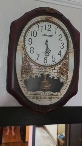 A wall clock with an oval face and a dark wooden frame is displayed. The clock face is white with black numbers and hands. The hands indicate approximately 12:57. Below the clock face is a decorative element resembling a fireplace with a pendulum swinging back and forth. The clock is mounted on a white wall, with a dark shelf or frame visible below it. The reflection in the dark surface shows a person's feet and legs, suggesting a person is standing in front of the clock. The overall environment appears to be an indoor space, possibly a room in a home.
