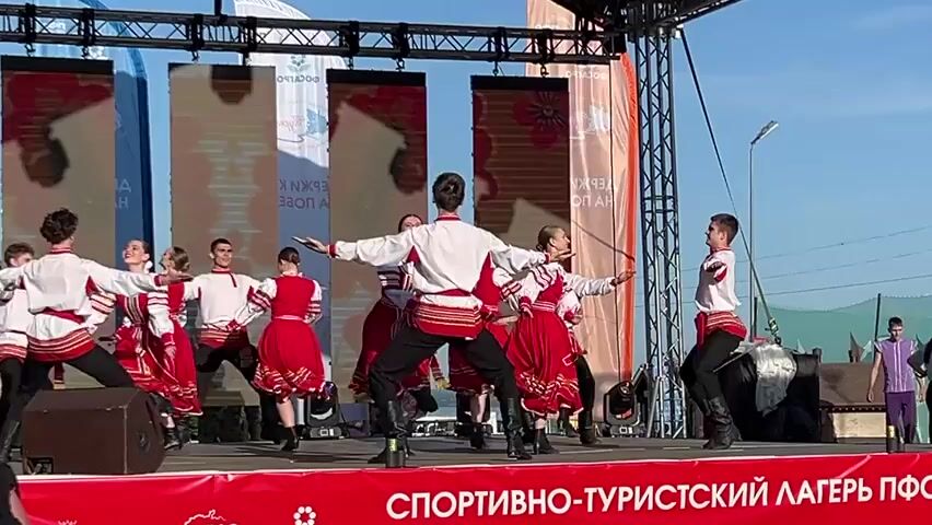 A group of young dancers performs a traditional Russian folk dance on an outdoor stage. The dancers, roughly equal in number of men and women, wear matching costumes of red, white, and black. The women's costumes include red skirts and white blouses with decorative trim, while the men wear black pants and white shirts with similar red trim, and all have black boots. The dancers move in unison, performing a series of coordinated steps, leaps, and turns. The stage has a red banner at the front with white text and logos. Backdrops with printed imagery and logos stand behind the performers. The sky is bright blue, and additional support structures and trees are visible behind the stage. A few indistinct individuals walk in the background to the right.