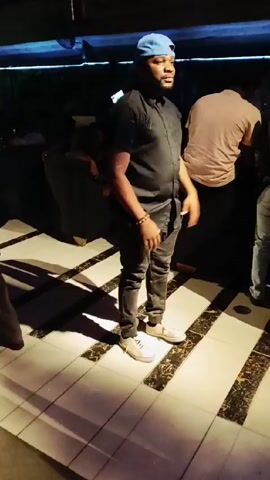 A man in a black shirt, grey jeans, and a blue baseball cap dances on a brightly lit tiled floor in a nightclub. He is surrounded by other people, some of whom are walking past. The lighting is low overall, but the dance floor is brightly lit. There are geometric patterns on the floor.