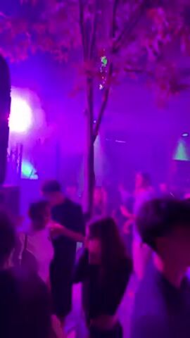Here is a detailed description of the video:

The video appears to capture a lively outdoor party or concert at night. The setting is vibrant with purple and green lighting, and there's a haze in the air, likely from a smoke machine. A tree is partially visible, its branches reaching into the lit space.

A large crowd of people is gathered, many wearing casual clothing. They appear to be enjoying themselves, possibly dancing or socializing. The camera focuses on various individuals within the crowd, including a person wearing a motorcycle helmet.

The overall atmosphere is energetic and festive, suggesting a vibrant nightlife event.