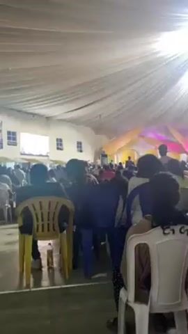 The video shows a large crowd of people gathered in what appears to be an indoor event space, possibly a church or event hall. The room is lit by bright overhead lights suspended from the ceiling, which is covered in white drapery giving it a tent-like appearance. A stage with yellow and pink decorations is visible at the far end of the room. The crowd is mostly standing and facing the stage, and there seems to be an event in progress. A metal lighting rig stands in the middle of the room. Many people are wearing casual clothing.