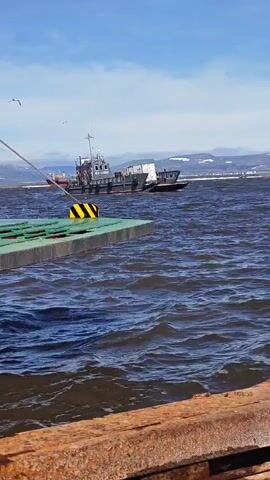 A tugboat with a gray cabin and dark hull navigates choppy, dark blue water. The tugboat is towing a white rectangular object. The tugboat is being pulled by a cable attached to a green platform with yellow and black striped buoys. The platform is in the foreground, obscuring part of the tugboat. The water is wavy, and there are mountains in the background. The sky is blue with scattered white clouds. Two birds are flying in the sky. The scene appears to be a harbor or bay.