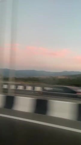 The video shows a view from a moving vehicle on a highway at dusk. The sky is a soft gradient of blue and pink, with scattered clouds. In the distance, rolling hills covered in trees are visible. A guardrail with alternating black and white stripes separates the highway from the landscape. Several cars are seen driving on the highway. One car, a dark blue SUV, is in the foreground, and another car is behind it. The overall impression is of a scenic drive through the countryside as the day ends.