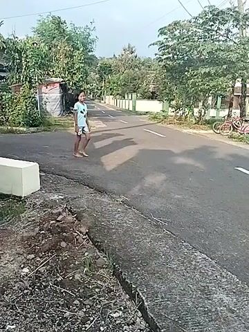 The video depicts a daytime scene on a paved road in what appears to be a residential or rural area of Bangsri, Indonesia. The weather is clear and sunny, as evidenced by the distinct shadows cast on the asphalt.

The primary subjects are two young girls. The first girl, with shoulder-length dark hair, wears a light blue t-shirt with a graphic (possibly a cartoon character) and patterned shorts. She is barefoot and walks leisurely across the road from left to right, looking towards the camera and smiling.

Following her, a second young girl, with dark hair tied back, wearing a light-colored tank top and dark leggings, runs barefoot across the road in the same direction. She also smiles broadly as she runs.

The setting includes a two-lane asphalt road with a solid white line marking one side. Both sides of the road are lined with abundant green trees. In the background, there are power poles with overhead lines, and what appear to be houses or buildings. A bicycle is visible parked on the grassy shoulder of the road on the right side of the frame. On the immediate left foreground, there's a patch of dirt and some concrete blocks. There is no visible text in the video.
