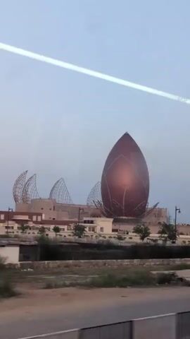 The video shows a large, tulip-shaped building with a copper-colored exterior and decorative white accents. The building is surrounded by other structures, some with pointed, cage-like roofs. In the foreground, there is a road with a few vehicles and some people walking and standing. The sky is overcast, and the overall impression is of an architectural landmark under construction or in an underdeveloped area.