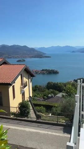 This video displays a panoramic view of a picturesque Italian island on a bright, clear day. The foreground features the corner of a yellow house with a terracotta roof, a white railing, and some greenery. The middle ground reveals a densely populated island with buildings featuring terracotta roofs, nestled against the vibrant blue waters of a lake. Several boats are visible docked around the island and on the water. In the background, distant mountains rise majestically, their peaks softly defined against the clear blue sky. The camera slowly zooms in, offering a closer look at the island's architecture and the surrounding landscape.