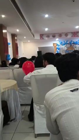 The video shows a dimly lit event hall with rows of chairs set up. A large red balloon floats near the ceiling. Several people are seated in the chairs, facing forward, suggesting they are attending an event such as a presentation or performance. In the background, a stage area is decorated with blue and white balloons and a silver curtain. Some people appear to be interacting or looking around. The overall atmosphere seems to be that of a formal gathering or celebration.