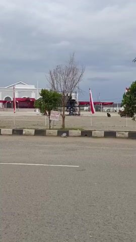 The video pans from left to right, showing an outdoor scene under a cloudy sky. In the foreground, there is an asphalt road with a white line. Beyond the road, there is a paved area with a low curb and a row of trees. In the background, there is a white building with red accents, a stage, and several red and white flags on poles. There are also some structures that appear to be tents or canopies. A person is visible sitting on a bench under a tree. There are also a few vehicles, including what looks like a motorcycle and a car, in the distance. The overall impression is a public gathering space decorated for an event.