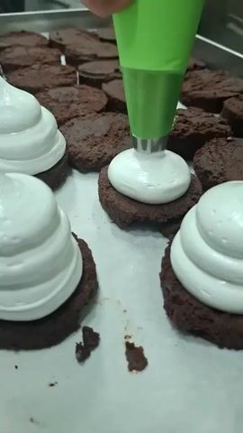 A gloved hand, holding a green piping bag with a silver tip, is decorating a tray of small, circular chocolate cookies with a thick, swirled topping of bright white frosting. A few cookies in the foreground are already decorated with the white topping, standing out against the darker chocolate base. The tray sits on a metal surface. The background shows more undecorated cookies, along with a glimpse of a commercial kitchen setting. The video focuses on the action of piping the frosting onto the cookies.