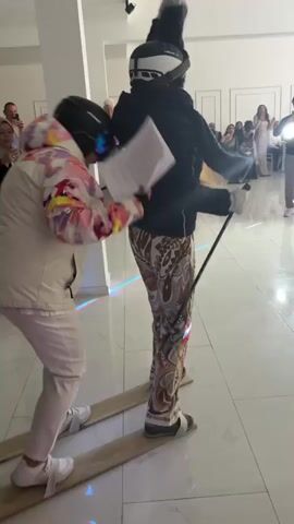 Two people are trying to walk on wooden skis indoors in front of a crowd of onlookers. The person in front is wearing a black jacket, printed pants, a ski helmet, and holding a ski pole and a stack of papers. The person behind is wearing a light-colored puffy jacket, white pants, and a ski helmet. They are on a shiny tiled floor in a large room, possibly a ballroom or event space. There are tables, chairs, and other people in the background. The person behind has their hands on the front person’s back, and they are struggling to move forward.