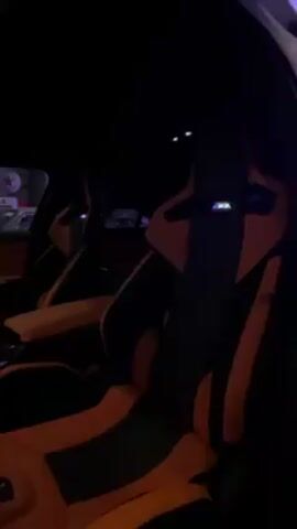 The video shows the interior of a car, specifically the driver's side. The seats are black with orange accents. The dashboard and door panels are also orange. There is a digital display on the dashboard, and the interior is illuminated with pink and purple ambient lighting. The car appears to be in a garage or similar indoor space with additional lighting visible.
