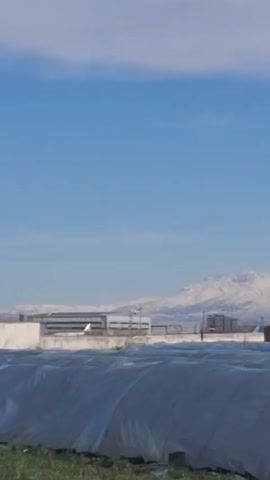 The video shows a daytime view of what appears to be a rural or semi-rural area. The sky is blue with scattered clouds. In the foreground, there are structures covered with white tarps. Behind them, there are buildings, including houses and what seems to be a larger industrial structure. In the distance, there is a mountain range covered in snow. The camera pans slightly to the right, showing more of the buildings and covered structures.
