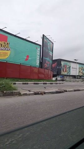 The video displays a roadside scene. Multiple billboards are visible, advertising products like Nescafé and Hennessy. Cars and a truck are on the road, suggesting traffic. People are seen near the billboards, one on a bicycle and another pushing a cart. A crane is also present in the background, indicating construction or industrial activity. The sky is overcast.
