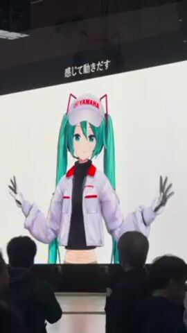 The video shows a presentation at a Yamaha event. The screen displays an animated Hatsune Miku character wearing a Yamaha-branded outfit, and then the Yamaha logo with the slogan "Revs Your Heart." There is a crowd of people watching the presentation. The event appears to be indoors, with spotlights and a high ceiling visible. Some attendees are looking at pamphlets. There is a wheelchair in the foreground.