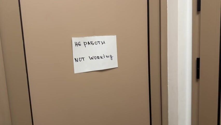 The video shows a closed door with a sign saying "не работает" (not working) in Russian and English. A person comes into view and removes the sign from the door. They then attach two new signs to the door, one with a male figure in a hat and the other with a female figure, indicating the door leads to a bathroom. The camera then focuses on the door with the new signs attached.