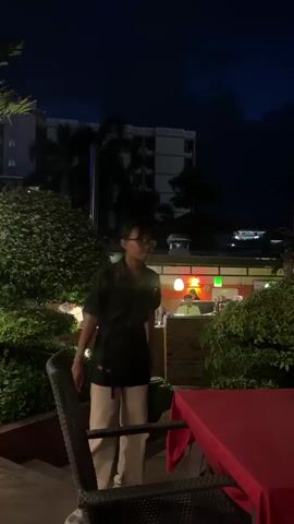 The video captures an outdoor dining scene at night, likely in Chiang Mai, Thailand. In the foreground, a table covered with a vibrant red tablecloth is set with a wicker chair. To the left, a woman wearing glasses, a black uniform shirt with red trim, and light-colored pants is walking, appearing to be a staff member. She walks on a lower level, with stairs visible behind her.

The background reveals a restaurant interior or covered area, illuminated by warm, colorful lights. Several individuals are visible working inside. One man, wearing a light-colored cap and a collared shirt, is the most prominent figure in the kitchen area. Another person in a dark shirt stands to his left. The overall atmosphere suggests a lively dining establishment in the evening. Lush green foliage surrounds the dining area, adding to the ambiance. The sky above is dark, indicating it is nighttime, and the silhouette of a building and palm trees can be seen against the night sky, hinting at an urban or semi-urban setting. The lighting from the restaurant creates a warm and inviting glow, contrasting with the dark night.