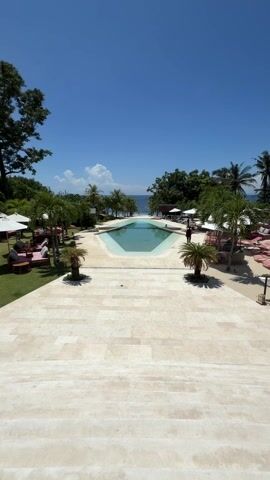 A sunny day illuminates an opulent resort. The view is from an elevated position, like the top of stairs, and the frame centers a geometric pool. The pool's light blue water reflects the clear sky above. Tall palm trees flank the pool on either side, adding to the tropical ambiance. On either side of the pool are lounge areas with red lounge chairs and white umbrellas. To the left, a large tree provides shade to the grass. A person dressed in black walks past the pool. The ocean stretches in the background. The paved area in the foreground appears to be made of large, light-colored stone tiles, giving a sense of space and luxury.