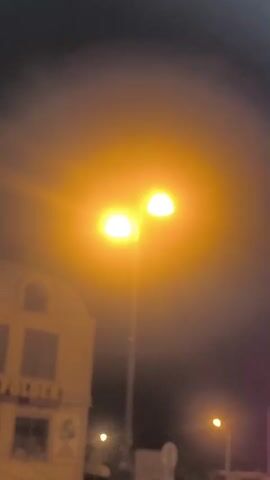 The video captures a street scene at night, illuminated by bright streetlights. In the foreground, lush green foliage is visible. A two-story building with a sign reading "MY FOLDER" is prominent on the left side of the frame. The building appears to be a commercial establishment, with an entrance and windows. A lamppost with two bright lights stands in the middle of the street. Further down the street, another lamppost and a white truck are visible. A black SUV drives across the scene from left to right. A-frame signs are placed on the sidewalk. The sky is dark, and a hazy glow emanates from the streetlights, suggesting the presence of fog or mist.