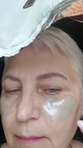 The video shows a close-up of a woman's face as she applies under-eye patches. Her blonde hair is visible, and she is wearing eye makeup. The patches appear to be clear or light-colored and are being applied to the skin beneath her eyes. Her right hand is visible, and her index finger is gently pressing one of the patches into place. The background is blurred, but a patterned fabric is visible at the top of the frame. The lighting is somewhat dim.