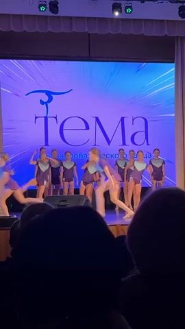 The video shows a group of young female dancers performing on a stage in front of an audience. The girls are wearing matching purple and navy athletic shorts and short-sleeved shirts. They are performing synchronized leaps and poses. In the background is a large screen displaying the name "Tema" in stylized lettering. The stage is lit with bright spotlights. The foreground shows the heads of audience members, partially obscuring the view.
