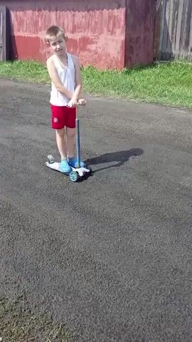 A young boy stands on a scooter outdoors on a paved surface. He is wearing a white tank top and red shorts. He holds onto the handlebars of the scooter, which is white and blue. In the background, there is a reddish-brown building and a wooden fence. Green grass is visible on the sides of the paved surface. The boy is looking at the camera. He shifts his weight and makes a facial expression. He then looks down at the scooter.