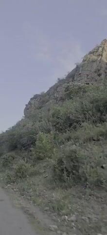 The video shows a narrow, unpaved road winding through a vegetated hillside. The road is on the left side of the frame, and the hillside is on the right. The hillside is covered in dense green bushes and trees, with some exposed rock formations at the top. The sky is a pale blue with some wispy clouds. The camera is moving forward, indicating that the video is likely taken from a vehicle. The overall impression is one of a remote, natural landscape.