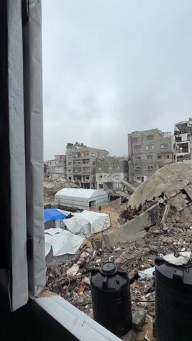 The video shows a scene of devastation, likely caused by conflict or disaster. Multiple buildings are heavily damaged, some reduced to rubble. In the foreground, several white tents are visible, with one blue tarp. The presence of these tents suggests they are serving as temporary shelters for displaced people. Two large, black water tanks are positioned in the foreground near the window from which the scene is being viewed. A single person walks through the area. The sky is overcast, contributing to the somber and bleak atmosphere.