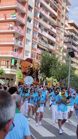 A parade is taking place on a city street. The street is lined with buildings, some of which have balconies adorned with flags. People are watching the parade from the balconies and sidewalks. In the foreground, a marching band wearing blue shirts and white pants is playing instruments, including saxophones and a sousaphone. Behind the band is a large, colorful float shaped like a bull with floral decorations. The float is adorned with flowers and other materials. The people in the band appear to be moving and performing. The overall atmosphere seems festive and celebratory.