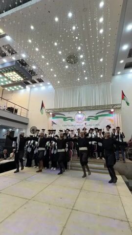 A group of men in traditional black clothing and headscarves are performing a dance on a raised stage. They are holding hands and moving in unison. Behind them, a group of graduates wearing black academic gowns and caps are standing. Two Palestinian flags are visible in the background, one on each side of the stage. The stage is lit with colorful lights, and a banner with Arabic writing is displayed above the graduates. The overall atmosphere appears to be a graduation ceremony or celebration.